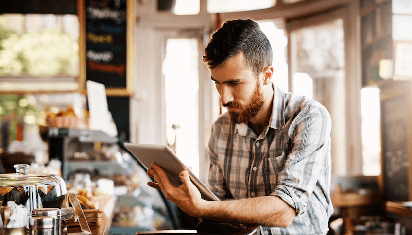 A man with a beard reads a tablet in a cafe, surrounded by pastries and a chalkboard menu.