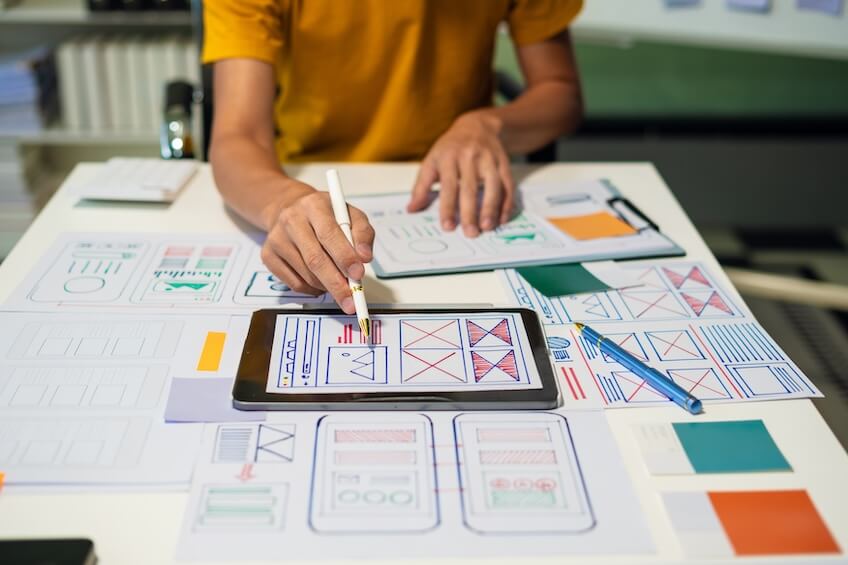 Person designing a website interface on a tablet with a stylus, surrounded by paper sketches and design layouts.