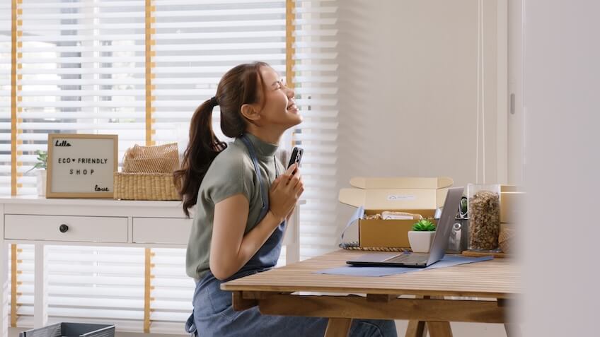Woman sitting at a table, smiling with eyes closed, holding a pen. Sign reads "Little ECO-FRIENDLY SHOP."