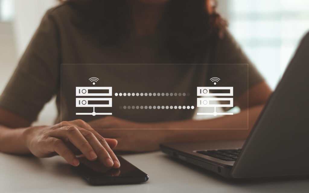 Person using a smartphone and laptop with a digital data transfer icon overlay, symbolizing wireless communication.