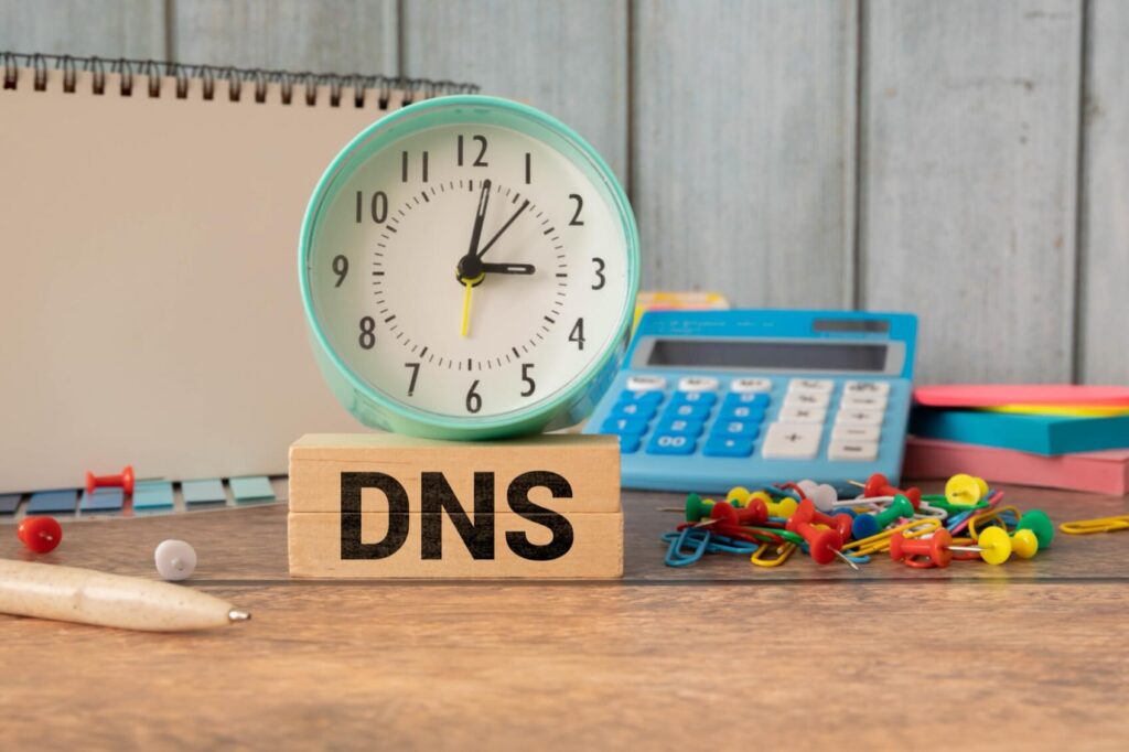 Wooden blocks with "DNS" text, an analog clock, calculator, and office supplies on a wooden desk.